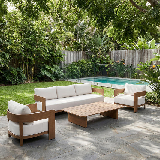 Outdoor lounge setting with cushioned sofa and armchairs featuring wooden frames, arranged around a low table on a patio beside a pool and garden