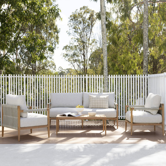Outdoor lounge set with light cushions and wooden frames, including a sofa, armchairs, and a coffee table on a patio with a white fence and trees in the background